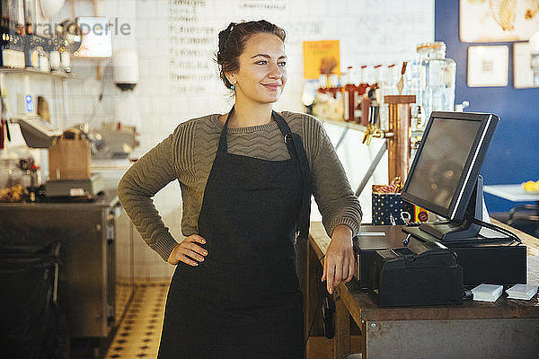 Selbstbewusste junge Besitzerin steht mit der Hand auf der Hüfte an der Kasse an der Kasse im Cafe