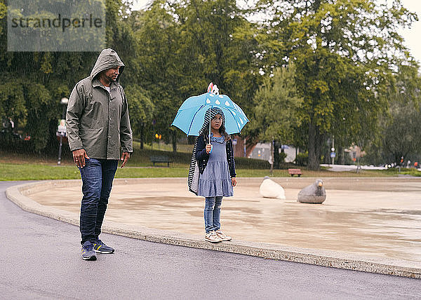 Vater sieht Tochter mit Regenschirm in voller Länge an  während er bei Regen im Park steht