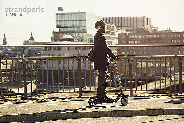Junger Mann fährt in voller Länge auf einem elektrischen Roller auf einer Brücke in der Stadt