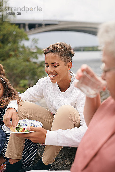 Lächelnder Teenager beim Essen  während er am Seeufer mit seiner Familie im Park sitzt