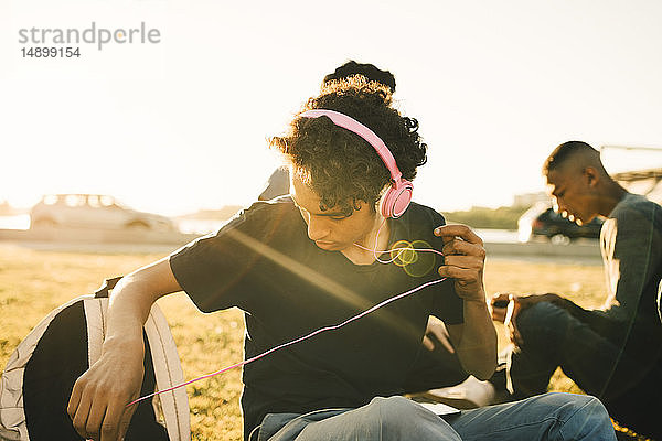 Teenager Junge hört Musik über Kopfhörer  während er an einem sonnigen Tag mit einem Freund zusammensitzt
