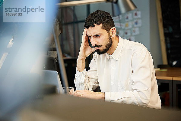 Angespannter Geschäftsmann schaut auf Laptop  während er im Kreativbüro sitzt