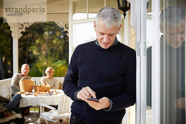 Reifer Mann benutzt Mobiltelefon  während er auf der Veranda mit Freunden im Hintergrund steht