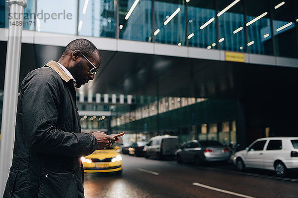 Seitenansicht eines Geschäftsmannes  der ein Smartphone benutzt  während er auf der Straße gegen ein Gebäude in der Stadt steht