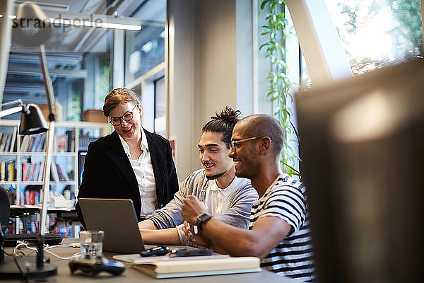 Männliche Unternehmer lächeln  während sie einer Bankmanagerin am Laptop am Schreibtisch im Kreativbüro erklären