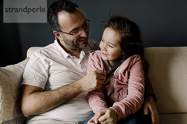 Lächelnder Vater sieht Tochter an  während er im Wohnzimmer auf der Couch sitzt