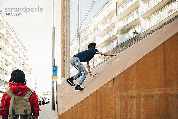 Sorgloser Teenager klettert an der Wand  während ein Freund am Bürgersteig in der Stadt steht