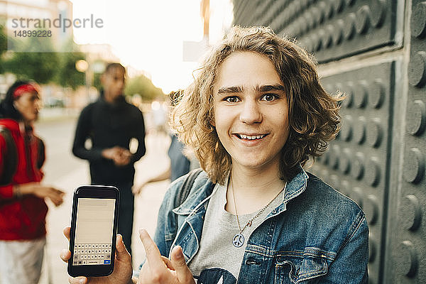 Porträt eines lächelnden jungen Mannes  der ein Mobiltelefon zeigt  während Freunde im Hintergrund auf der Straße stehen