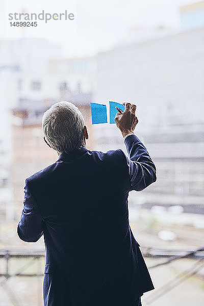 Rückansicht eines Geschäftsmannes  der während der Strategieplanung im Kreativbüro Haftnotizen auf Fensterglas klebt