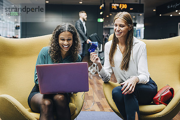Lächelnde multiethnische Kolleginnen diskutieren über Laptop und Kreditkarte  während sie in der Flughafen-Lobby sitzen