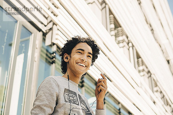 Niederwinkelansicht eines fröhlichen Teenagers  der über Kopfhörer Musik hört  gegen ein Gebäude in der Stadt