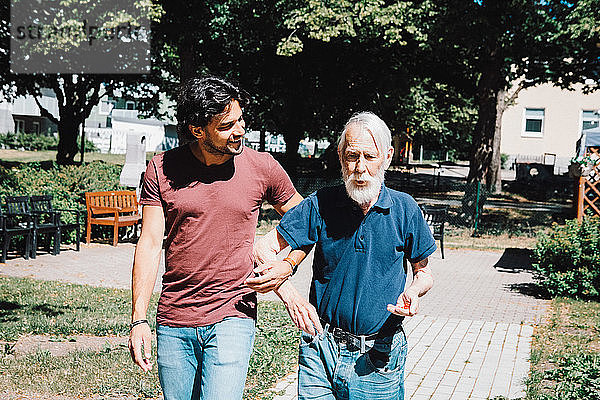 Lächelnder männlicher Pfleger schaut  während er Arm in Arm mit einem älteren Mann im Pflegeheim geht