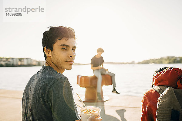 Porträt eines jungen Mannes  der an einem sonnigen Tag auf der Promenade sitzt und eine Mahlzeit in einem Behälter hält