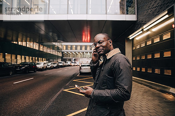 Porträt eines Geschäftsmannes mit Smartphone  der Musik hört  während er auf der Straße gegen Gebäude in der Stadt steht
