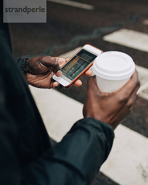 Mitschnitt eines Geschäftsmannes mit Kaffee mit Mobiltelefon  der auf einem Zebrastreifen in der Stadt steht