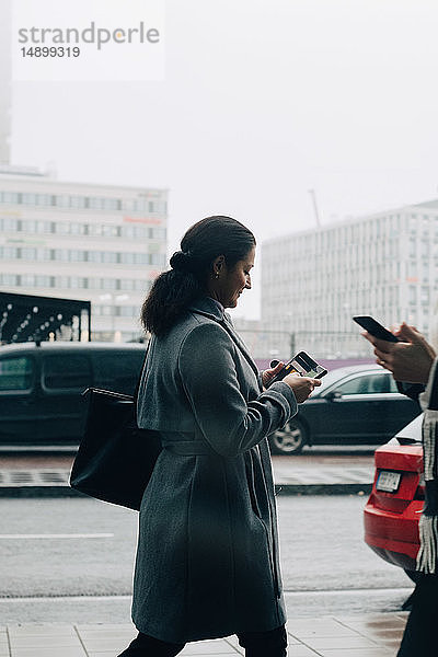 Geschäftsfrauen benutzen Mobiltelefone beim Gehen auf dem Bürgersteig gegen den Himmel in der Stadt