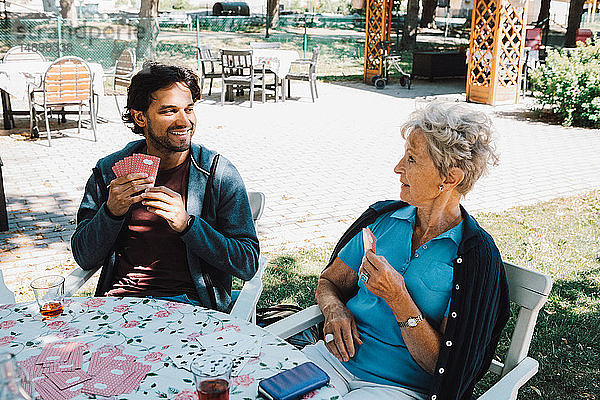 Ältere Frau im Ruhestand spielt Karten mit einem lächelnden männlichen Hausmeister  der am Tisch im Hinterhof sitzt