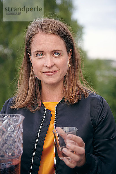 Porträt einer lächelnden jungen Frau  die während einer Party auf der Terrasse ein Getränk in der Hand hält