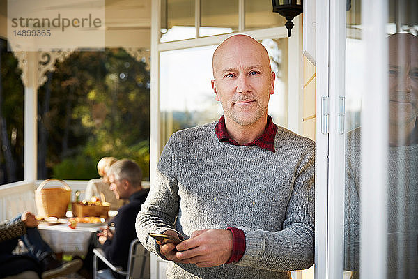 Porträt eines selbstbewussten  kahlköpfigen  reifen Mannes  der auf einer Veranda mit Freunden im Hintergrund steht