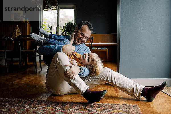 In voller Länge ein glücklicher  verspielter Vater  der seine Tochter trägt  während er zu Hause auf einem Parkettboden sitzt