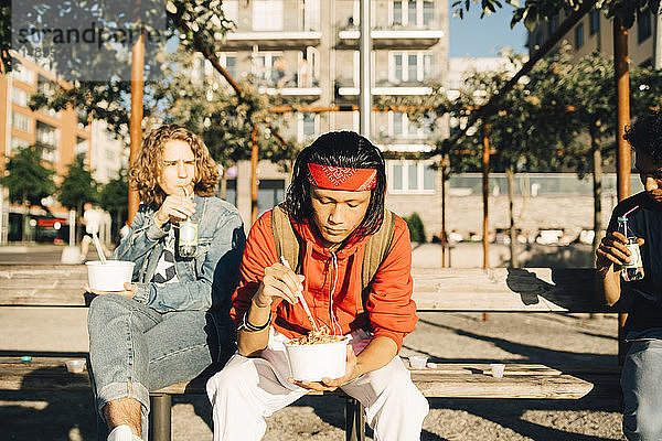 Freunde essen aus Containern  während sie an sonnigen Tagen in der Stadt auf einer Bank sitzen
