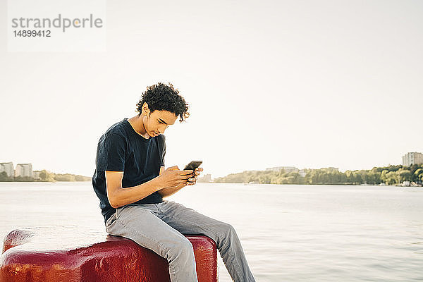 Teenager  der ein Mobiltelefon benutzt  während er auf einem Poller am Fluss vor klarem Himmel am sonnigen Tag sitzt