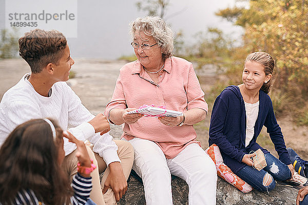 Glückliche Großmutter schenkt Enkel  während sie mit Enkelinnen im Park beim Picknick sitzt