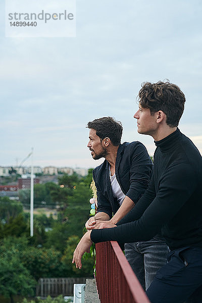 Männliche Freunde schauen weg  während sie am Geländer auf der Terrasse gegen den Himmel stehen