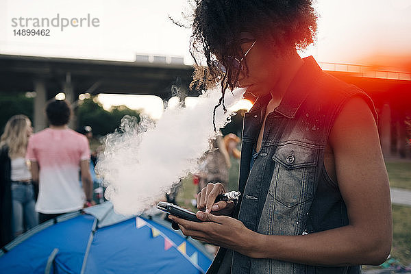 Seitenansicht eines jungen Mannes  der raucht  während er bei einer Musikveranstaltung ein Mobiltelefon benutzt