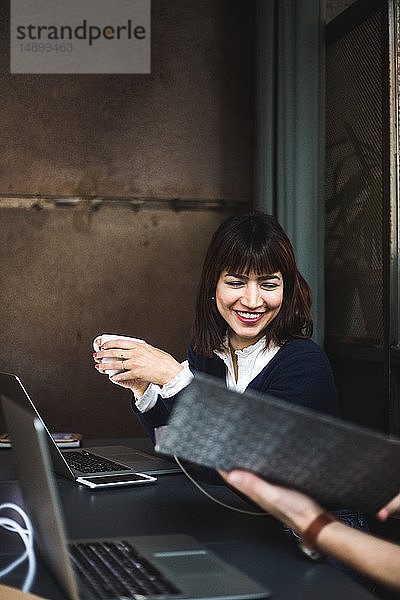 Lächelnde Unternehmerin hält Kaffeetasse in der Hand und diskutiert mit Kollegin über Dokument am Schreibtisch im Büro