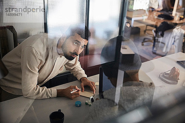Geschäftsmann schreibt im Notizblock  während er mit einem männlichen Kollegen am Schreibtisch im Kreativbüro diskutiert