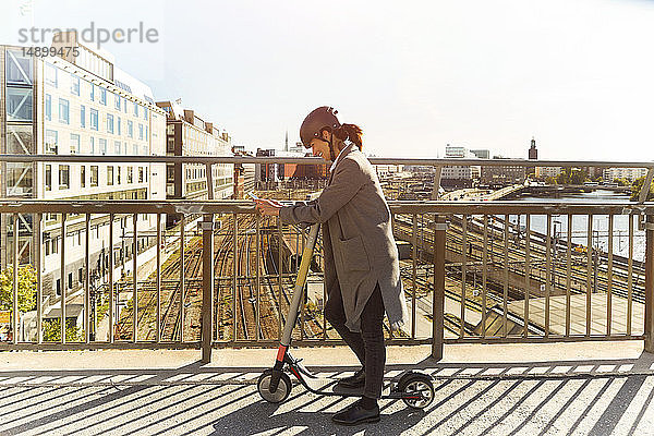 Seitenansicht in voller Länge einer reifen Frau  die ein Smartphone benutzt  während sie mit einem elektrischen Roller auf einer Brücke in der Stadt steht