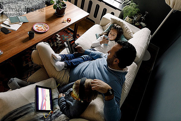 Hochwinkelansicht von Vater und Töchtern unter Verwendung verschiedener Technologien auf der Couch im heimischen Wohnzimmer