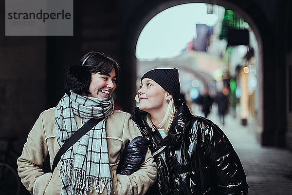 Lächelnde junge Freundinnen  die einander von Angesicht zu Angesicht gegenüberstehen  während sie im Winter auf der Straße in der Stadt stehen