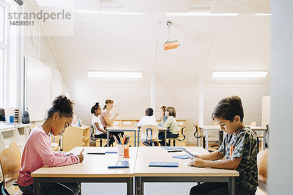 Junge und Mädchen schreiben am Schreibtisch  während Freunde mit dem Schüler im Hintergrund lernen