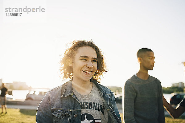 Lächelnder junger Mann  der wegschaut  während sein Freund im Hintergrund vor dem klaren Himmel an einem sonnigen Tag steht