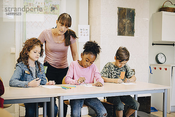 Lehrer betrachtet Schüler  die in der Grundschule am Schreibtisch schreiben