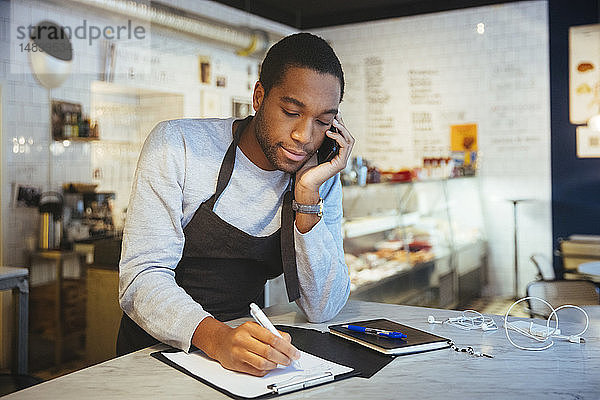 Selbstbewusster junger männlicher Angestellter spricht durch ein Smartphone  während er in einem Feinkostladen an der Zwischenablage schreibt