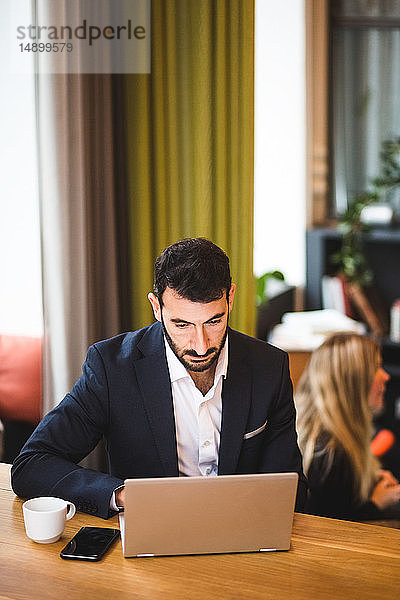Selbstbewusster männlicher Unternehmer benutzt Laptop  während er im Büro am Tisch sitzt