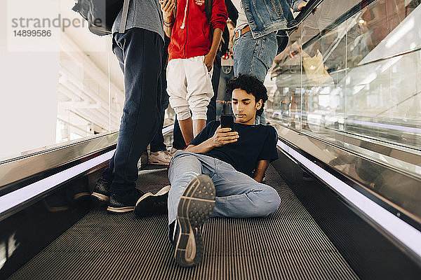 Teenager  der ein Mobiltelefon benutzt  während er bei Freunden auf dem Laufsteg eines Einkaufszentrums sitzt