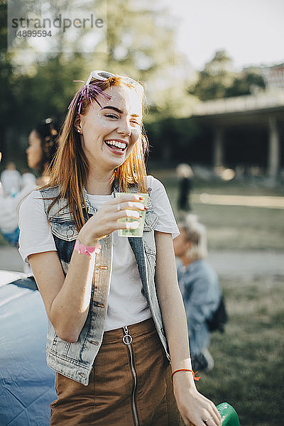 Fröhliche Frau trinkt im Stehen auf Rasen beim Musikfestival