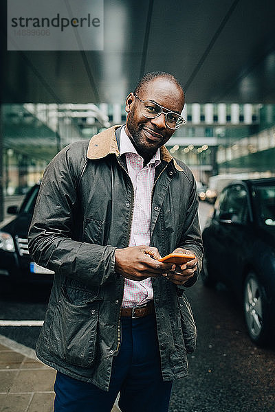 Porträt eines Geschäftsmannes  der ein Smartphone benutzt  während er auf der Straße gegen Gebäude in der Stadt steht