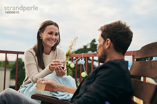 Fröhliche Frau unterhält sich mit einem männlichen Freund  während sie während des geselligen Beisammenseins auf der Terrasse ein Getränk hält