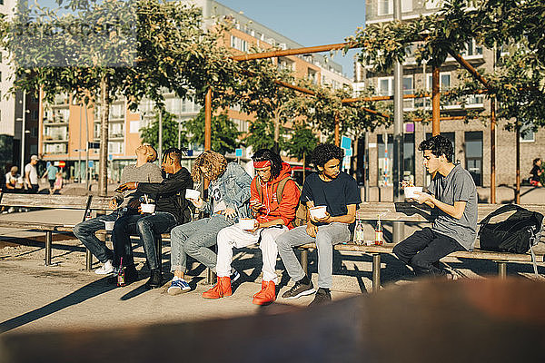 Männliche Freunde  die essen  nehmen Essen heraus  während sie nebeneinander auf einer Bank in der Stadt sitzen