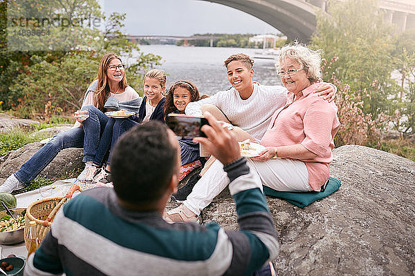Mann fotografiert glückliche Familie beim Essen auf einem Felsen im Park während eines Picknicks