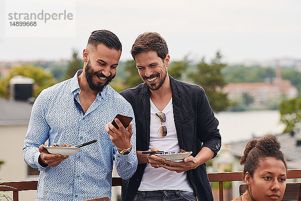 Lächelnder Mann zeigt einem Freund sein Handy  während er während der Party auf der Terrasse ein Essen hält