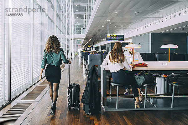 Rückansicht einer Geschäftsfrau in voller Länge  die mit ihrem Gepäck von Kollegen  die im Abflugbereich des Flughafens warten