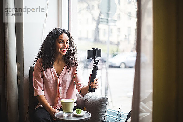 Selbstbewusste junge einflussreiche Frau  die am Fenster sitzend am Fenster eines kreativen Büros durch ein Mobiltelefon auf einem Stativ vloggt