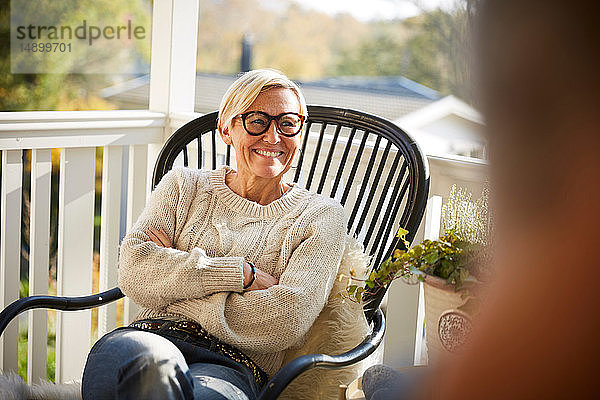 Fröhliche reife Frau mit verschränkten Armen  die auf der Veranda sitzend wegschaut