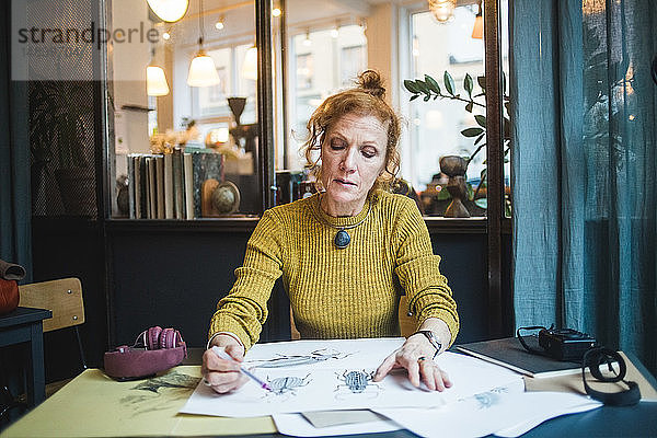 Selbstbewusste reife Illustratorin zeichnet auf Papier am Schreibtisch im Kreativbüro